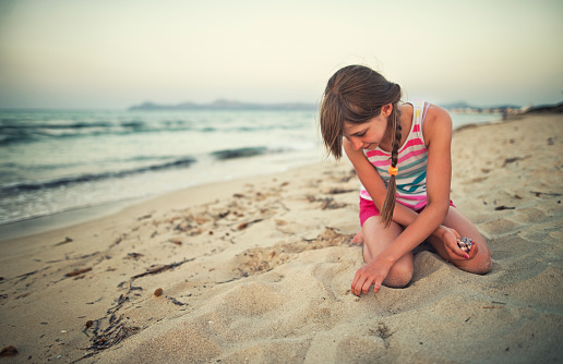Little girl on the beach is picking up sea shells from the sand. The girl aged 10 is smiling and kneeling on sand. She holds a handful of shells in one hand and is picking up another sea shell with the other hand.