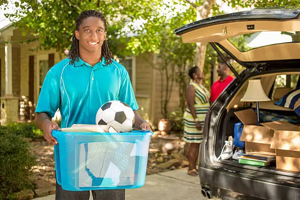 African descent boy heads off to college or moves away from home.  The 18-year-olds' parents are helping him pack up his car as he gets ready for the big move.  He is excited to start his college adventures. He carries a box of his belongings.  Family events.  Back to school.