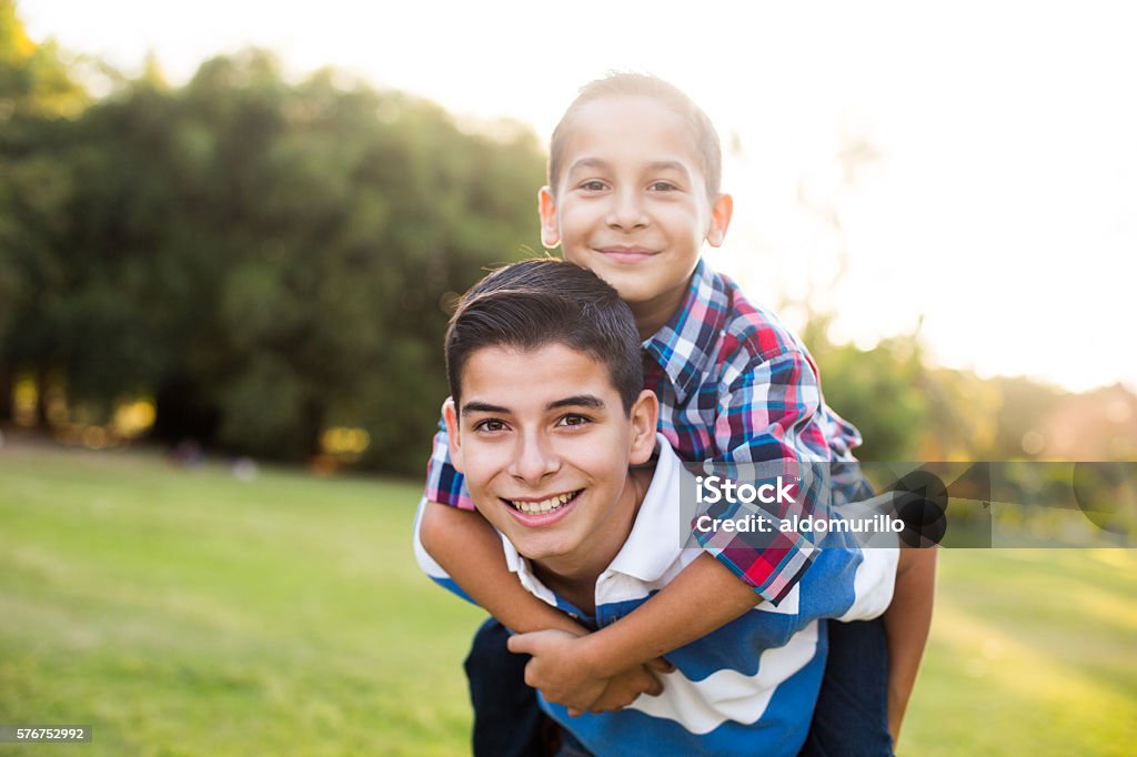 Teen boy carrying younger brother on back A teen boy carrying his younger brother on the back and smiling at the camera in a horizontal waist up shot outdoors. Teenager Stock Photo
