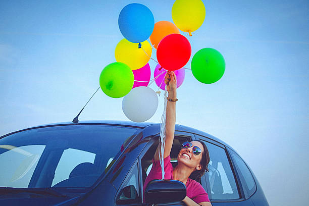 Cheerful woman holding colorful balloons, peeps out the car window stock photo