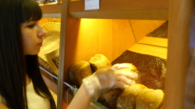 Woman chooses and putting a fresh bread loaf into a package in the supermarket. Young girl taking a loaf of bread from the shelf, smelling it and putting it into the basket. Shopping in the grocery