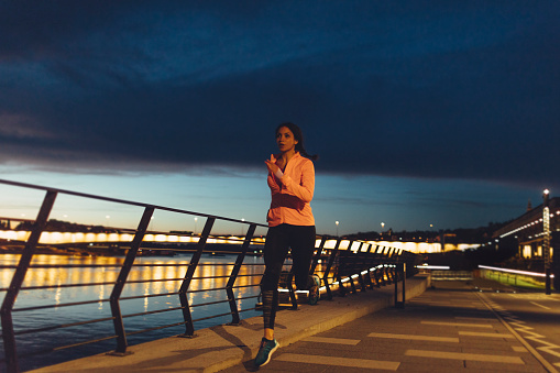Young woman jogging near river at night. She is wearing modern sporty clothing. With cityscape in background.