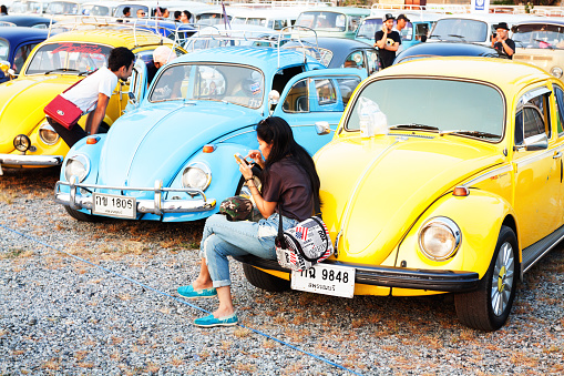 Bangkok, Thailand - February 15, 2014: A thai girl is sitting on engine bonnet of yellow VW Beetle oldtimer. Oldtimer is parked in a row of many VW Beetle oldtimers. Scene is on Siam VW festival at Fashion Island. Thai people are showig their cars and oldtimers. Woman is using with mobile. Between cars are some men.