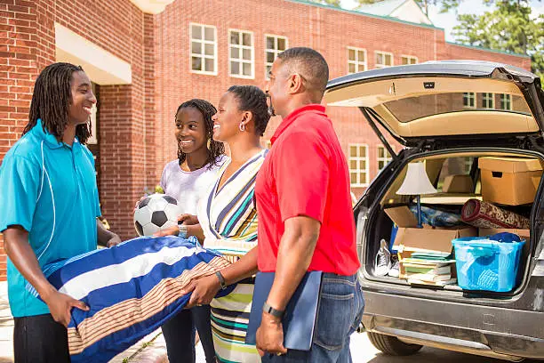 African descent boy heads off to college.  The 18-year-olds' parents and sister are all helping him unpack his car as he moves into the college campus dorm.  He is excited to start his school adventures. He  carries a pillow and textbooks.  Family events.  Back to school.
