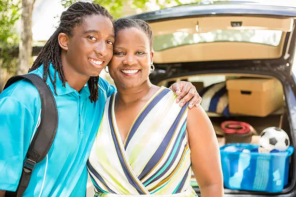 African descent boy heads off to college or moves away from home.  The 18-year-olds' mother is helping him pack up his car as he gets ready for the big move.  He is excited to start his college adventures and gives mom a big hug. He wears a backpack.  Family events.  Back to school.