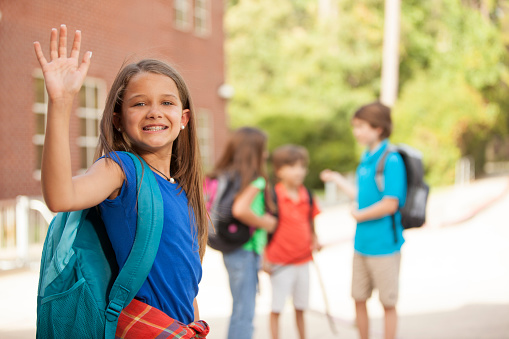 Multi-ethnic group of elementary-age students on elementary school campus.  One happy, Latin descent girl in foreground turns to wave goodbye to mom.  The friends in background all wear backpacks and are excited about going back to school.  Bright sunny autumn day.  Mixed-aged children.