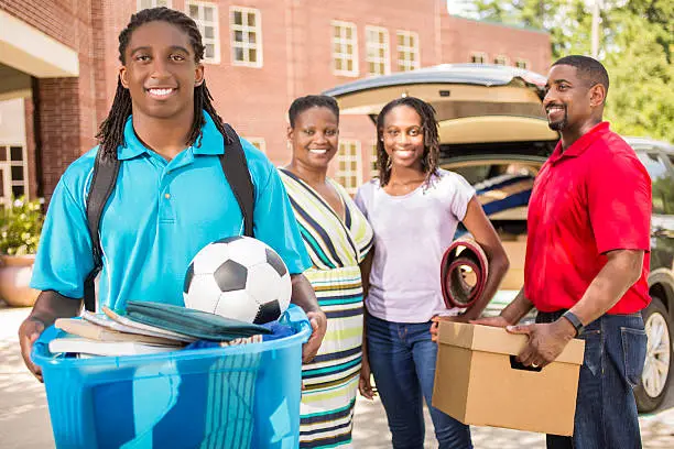 African descent boy heads off to college.  The 18-year-olds' parents and sister are all helping him unpack his car as he moves into the college campus dorm.  He is excited to start his school adventures. He  carries a backpack and a tub full of his belongings.  Family events.  Back to school.