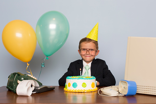 A young businessman with birthday cake is at an office birthday party. He is wearing a suit and tie, birthday hat, and a happy expression on his face. His business has just made it past the one year mark. Happy business anniversary.