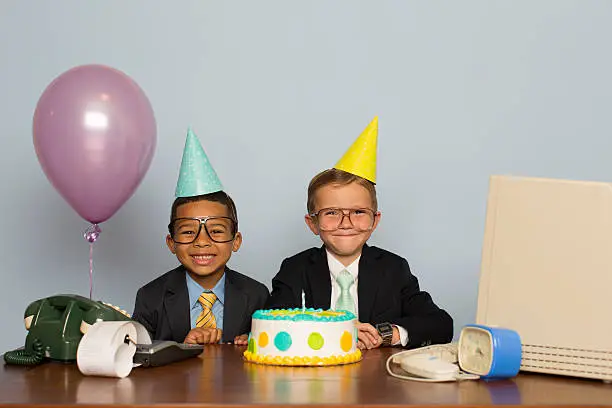 Two young businessmen celebrate their one year business anniversary with birthday cake are at an office birthday party. They are wearing a suits and ties, birthday hats, and have happy expression on their faces. They are sitting at a computer desk wearing party hats and having a fun time.   