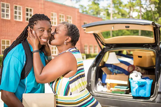 African descent boy heads off to college.  The 18-year-olds' mother is helping him unpack his car and says goodbye with a kiss as he moves into the college campus dorm.  He is excited to start his school adventures. He  carries backpack and textbooks.  Family events.  Back to school.