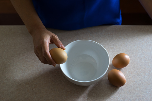 Asian female wearing blue apron holding brown chicken egg in her hand.