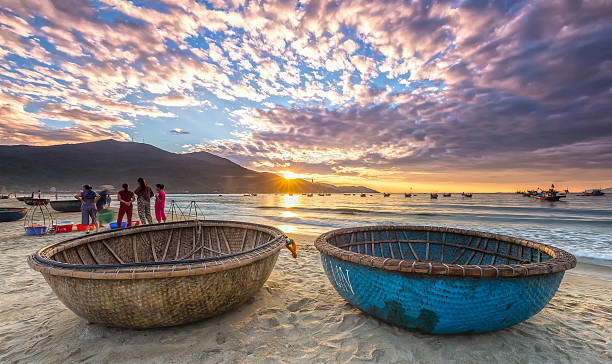 Sunrise over seaside Da Nang, Vietnam, June 26th, 2015: Sunrise over sea with sunlight to create stars and double pannier foreground boat, people exercising and watching fishermen bring fish on sale in Da Nang, Vietnam basket boat stock pictures, royalty-free photos & images