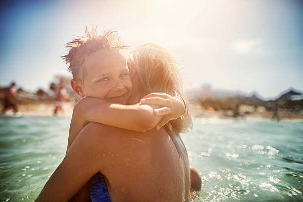 Smiling little boy and his mother are embracing in the beautiful sea. The litte boy is is holding to his mother and smiling to the camera. The sky is clear and the sun is shining. Shallow focus on the little boy.