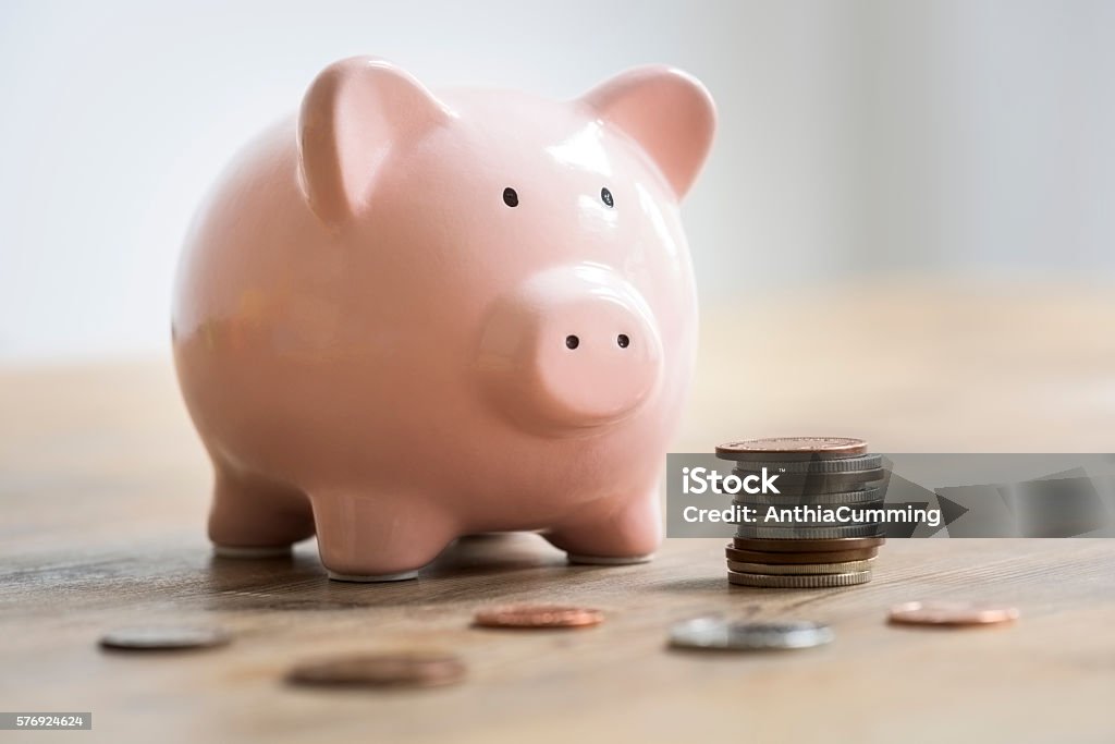 Coins beside a pink piggy bank to deposit and save Loose coins and a stack of coins are lying on a wooden table in front of a pink ceramic piggy bank. The selective focus is on the piggy banks face. The coins are bronze and silver and are to be deposited into the piggy bank as savings. Piggy Bank Stock Photo