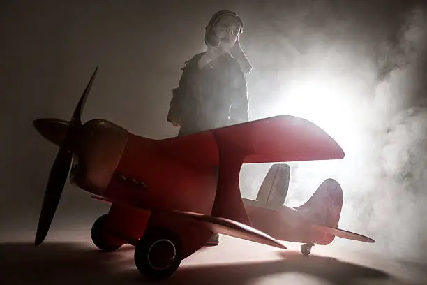 Cute little boy dressed in leather jacket and retro pilot helmet standing in a smoke. The wooden model of a red biplane on foreground. The little boy is screaming turning his head. He is looking away. Studio shooting with backlighting behind