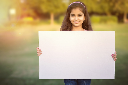 Smiling little asian girl holding an empty white banner in a park for you sample text
