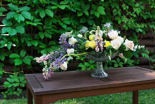 Beautiful flower composition outdoors. Wedding floristic decoration at wooden table, bouquet of colorful wildflowers and white roses in glass vase at green leaves background
