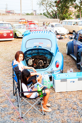 Bangkok, Thailand - February 15, 2014: Two thai women are sitting next to VW Beetle oldtimer with open engine bonnet. One girl is using tablet. Around and in background are more VW oldtimers and thai people. Scene is on annual public Siam VW festival.