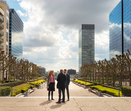 A team of three colleagues talking in the main thoroughfare of La Defense, the main business district of Paris, with the Arc de Triomphe visible in the distance.