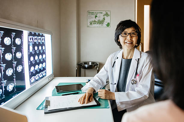 Female japanese doctor talking to a patient Female mature japanese doctor talking to a patient in her doctor's office. Illuminated only by MRI scans on her right hand side. japanese female doctor stock pictures, royalty-free photos & images