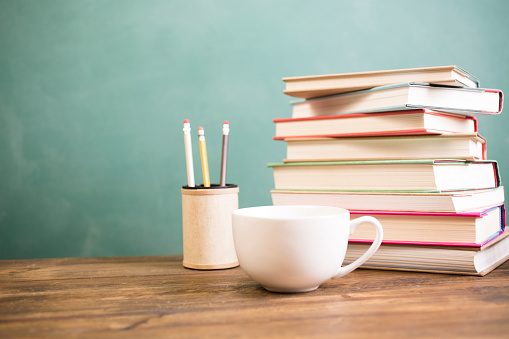 It's back to school time!  A large stack of teacher's books, pencils, and coffee cup at side makes frame composition.  The pile of objects lies on top of a wooden school desk with a green chalkboard in the background.  The blank blackboard in the background makes perfect copyspace!  Education background themes.  Teaching concept.