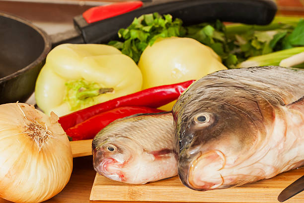 Two fresh raw river fishes, carp and crucian close up on a cutting board with onions, peppers, green herbs, red chili on table,next to frying pan, home cooking