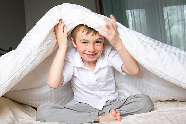 istock Smiling boy hiding in bed under a white blanket or 577341754