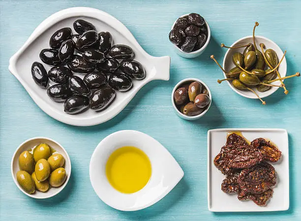 Mediterranean snack assortment. Black and green Greek olives, capers, olive oil and sun-dried tomatoes over turquoise blue background, top view