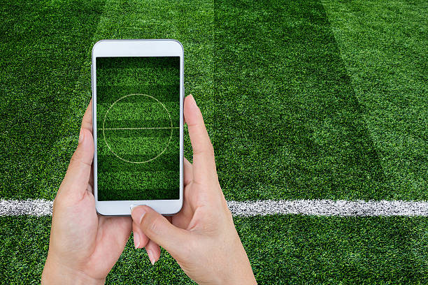 サッカースタジアムの画面で携帯電話を持って手。 - soccer soccer field grass american football ストックフォトと画像