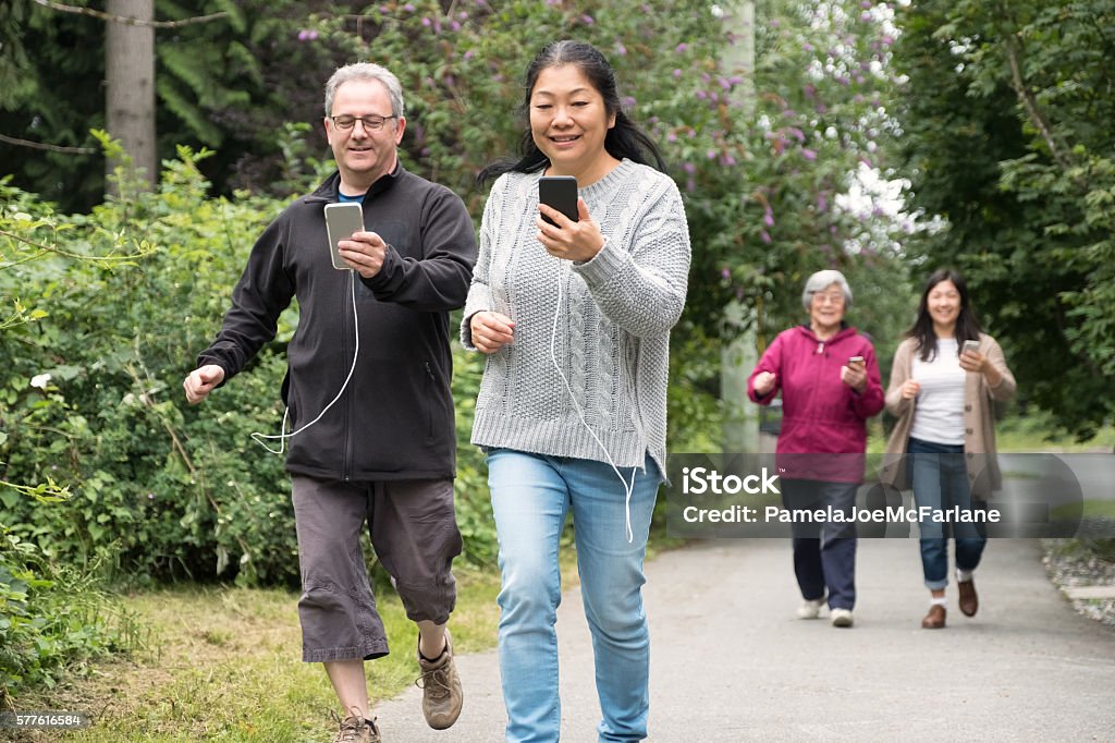 Mature Couple with Smartphones Run While Playing Augmented Reality Game Excited, mature interracial couple run ahead of their daughter and her grandmother while playing an augmented reality game on their smartphones.  The man and woman each have power cords attached to chargers in their pockets to give them longer battery life.  They are hurrying down a path in a public park in North Vancouver, British Columbia, Canada Senior Adult Stock Photo