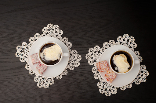 Two mugs of coffee with ice-cream, Turkish delight on a saucer, on white lace napkins. Top view, black background