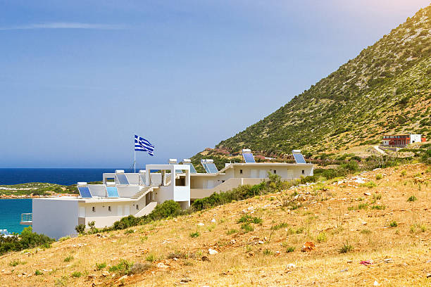 Modern Greek architecture - new white building in constructivist style, stands on shore of Cretan sea on roof mounted solar panels with hot water heaters, on flagpole the flag of Greece. Resort village Bali, Rethymno, Crete, Greece