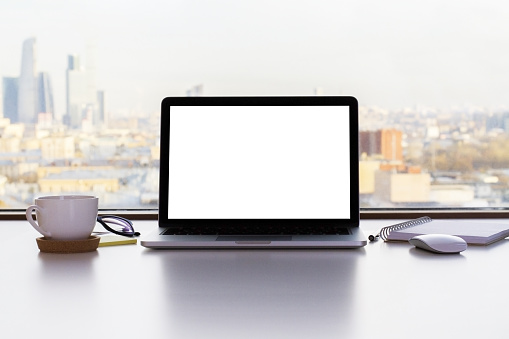 Front view of blank white laptop screen, coffee cup and other items on white desktop with sunlit city in the background. Mock up