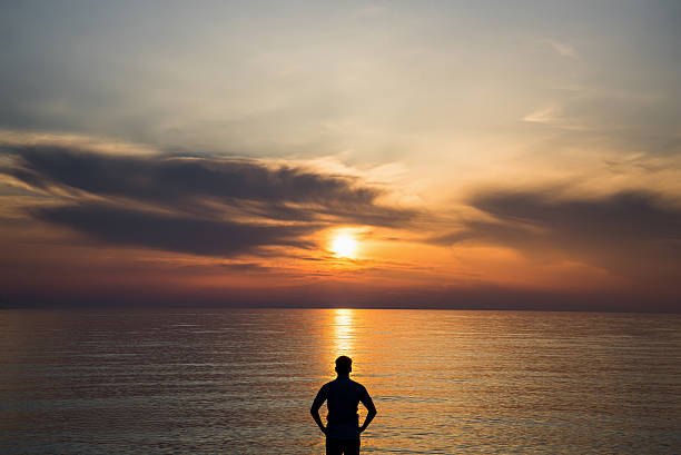 Young man standing at the beach in front of amazing sea view at sunset or sunrise and thinking about his future. Rear view.