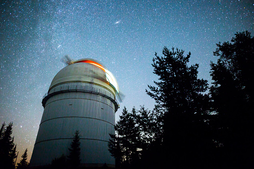 Rozhen astronomical observatory under the night sky stars. Blue sky with hundreds of stars of the Milky way. Observatory in a pine trees forest in the mountain.