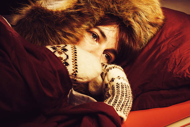 Woman lying in a bed wearing fur hat and mittens and freezing.