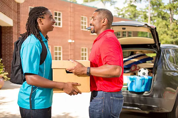 African descent boy heads off to college.  The 18-year-olds' father is helping him unpack his car and says goodbye as he moves into the college campus dorm.  He is excited to start his school adventures. He  carries backpack and box.  Family events.  Back to school.