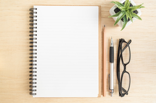 Desk above, Top view of open spiral notebook with brown pencil, pen, black eye glasses and small plant pot on wooden background