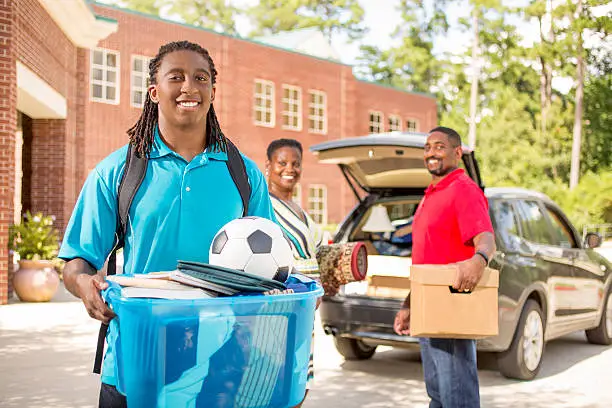 African descent boy heads off to college.  The 18-year-olds' parents are all helping him unpack his car as he moves into the college campus dorm.  He is excited to start his school adventures. He  carries a backpack and a tub full of his belongings.  Family events.  Back to school.
