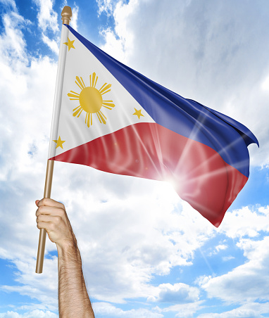 Person holding the national flag of the Philippines high in the air against a bright sky and sun rays.