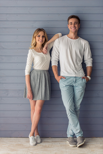 Full length portrait of beautiful young couple in casual clothes looking at camera and smiling, standing against gray wall