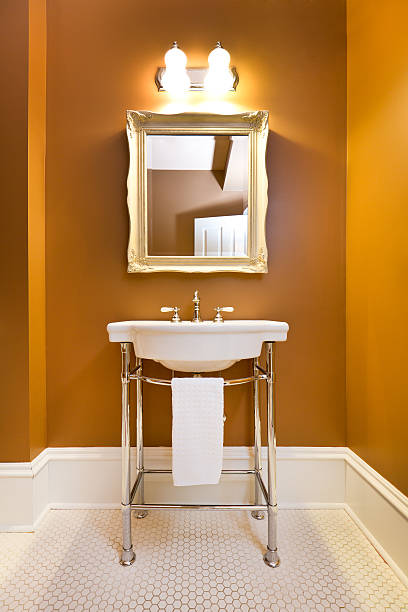 A modern contemporary classic bathroom design, furnished with a old fashion retro pedestal sink, classic lighting fixtures on the wall, classic framed mirror, old fashion white tile pattern on the floor. Photographed in vertical format.