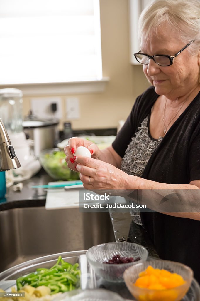 Real situation-Senior woman peeling a hard boiled egg in kitchen Real Situation- Caucasian senior woman is standing by the kitchen sink and peeling a hard boiled egg. The sink is stainless steel. There is a serving tray with an assortment of vegetables, fruit and eggs next to the sink. Shot with Canon 5D Mark 3. Peeling Food Stock Photo