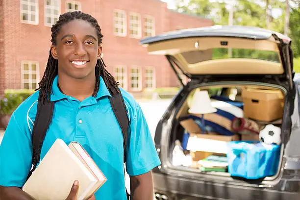 African descent boy heads off to college.  The 18-year-olds is unpacking his car as he moves into the college campus dorm.  He is excited to start his school adventures. He carries a backpack and books.  Back to school.