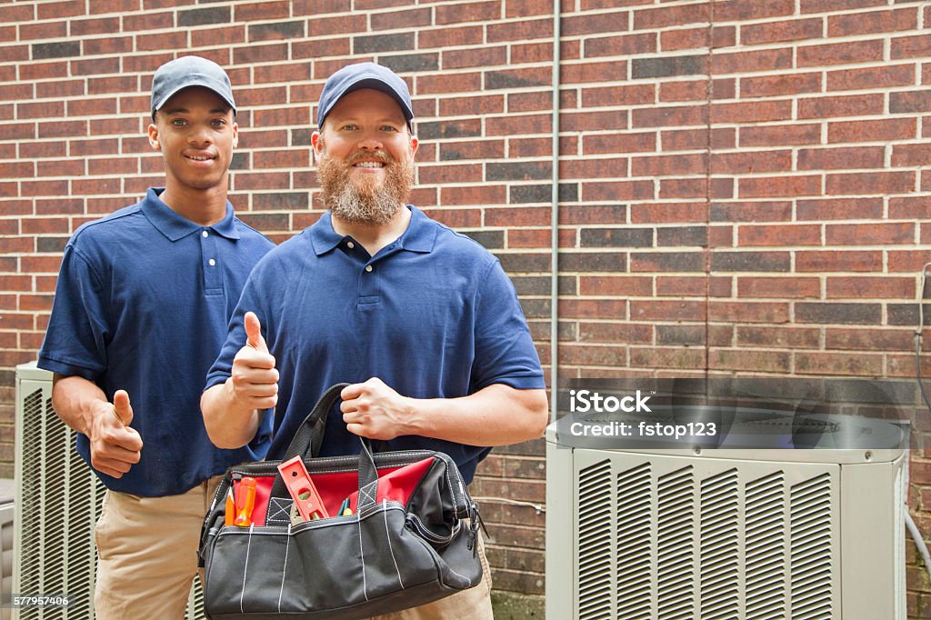 Air conditioner repairmen work on home unit.  Tool bag. Multi-ethnic team of men repairing a home's air conditioner unit outdoors. They have completed the repairs.  They both wear blue uniforms and man in foreground holds a tool bag.  Thumbs up for a job well done! Air Conditioner Stock Photo
