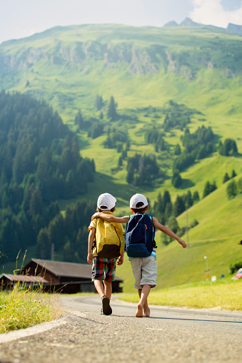 Two little children, boy brothers with backpacks travel on the road to scenic mountains, summertime