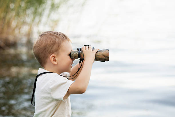 little boy looking through binoculars on river bank - searching child curiosity discovery imagens e fotografias de stock
