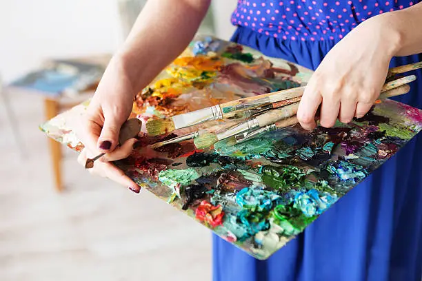 artists brushes and oil paints on wooden palette. macro artist's palette, texture mixed oil paints in different colors and saturation. palette with paintbrush and palette-knife in artistâs hands. The girl artist holding a palette with oil paints