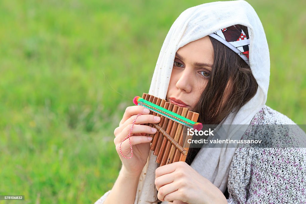 Young sad woman playing panpipe Sad young woman sitting on grassland and playing on panpipe. Looking down, with headband and scarf on head. Green grass on background. Focus on foreground. Panpipe Stock Photo