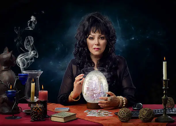 Portrait of stunning mature crystal gazer sitting at the desk surrounded by burning candles. Black haired female fortuneteller is scrying with big crystal egg and looks intently at the camera. Horizontal indoors shot on dark background